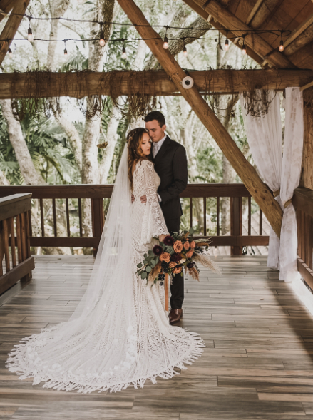 A bride in a long-sleeved lace wedding dress and a groom in a dark suit embrace on a covered wooden porch.