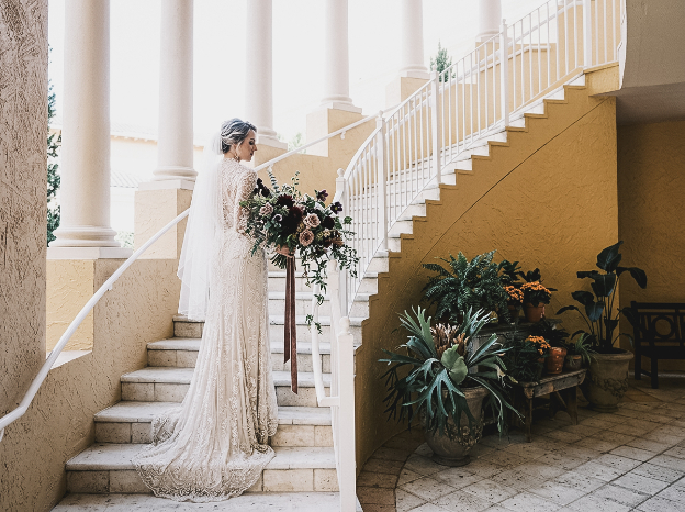 A bride in a long-sleeved lace wedding dress and veil stands on stone stairs, holding a large bouquet of dark flowers.