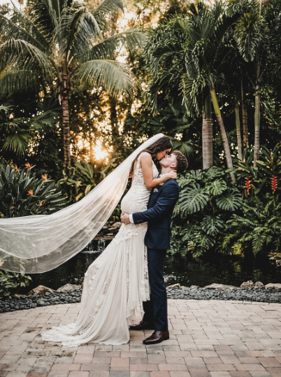 A bride in a long veil and gown kissing her groom who is lifting her up in front of a tropical garden.