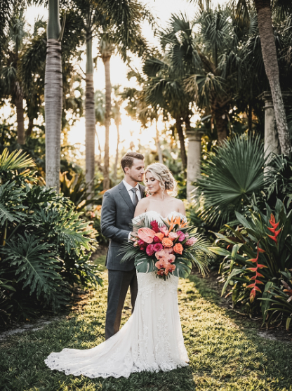 A bride in a white wedding gown and a groom in a grey suit stand together in a tropical garden, holding a large, colorful bouquet of exotic flowers.