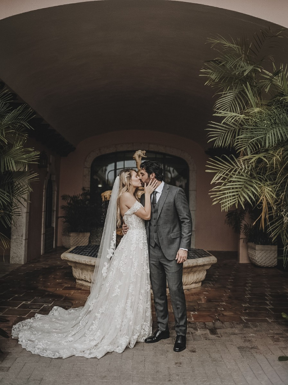 A bride in a lace wedding dress and a groom in a grey suit kissing in front of a stone fountain.