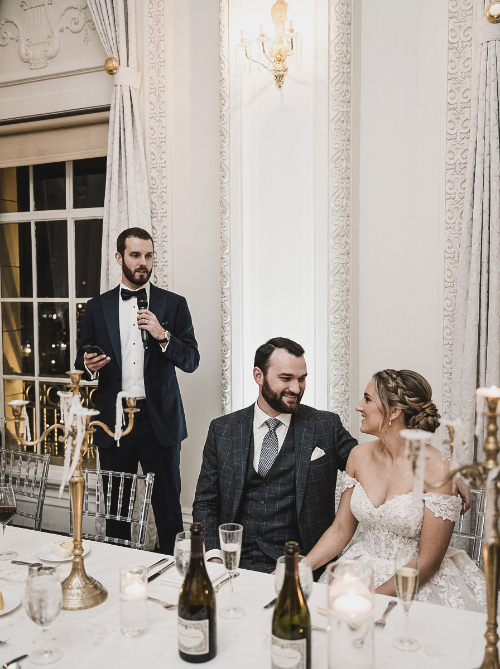 A man in a tuxedo stands and speaks into a microphone while a groom in a grey suit and a bride in a white wedding dress smile at each other at a reception table.