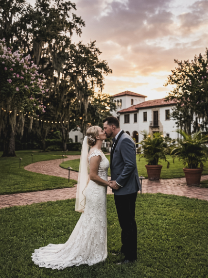 A bride and groom kissing on a lawn in front of a villa at sunset.