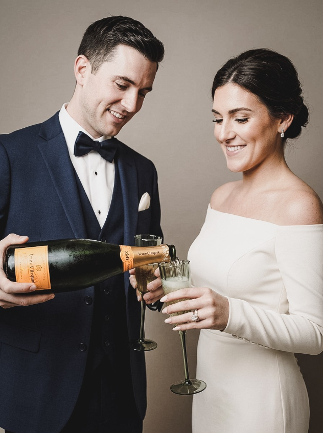 A man in a tuxedo pouring Veuve Clicquot champagne into a glass held by a woman in an off-the-shoulder white dress.