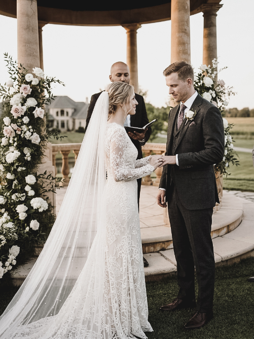 A bride and groom exchange rings during their outdoor wedding ceremony while an officiant stands between them.