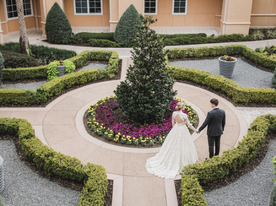 A bride in a long white gown and a groom in a dark suit hold hands while standing in a circular garden.