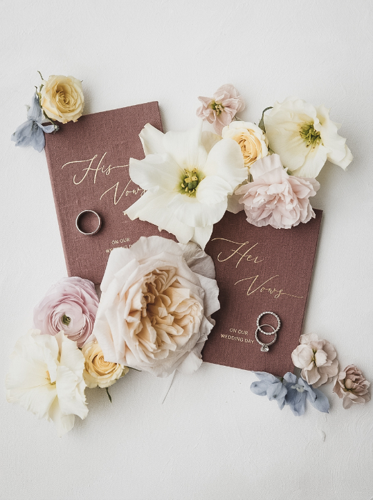 Two wedding vow booklets labeled “His Vows” and “Her Vows” surrounded by white and pink flowers, with wedding bands placed on each booklet.