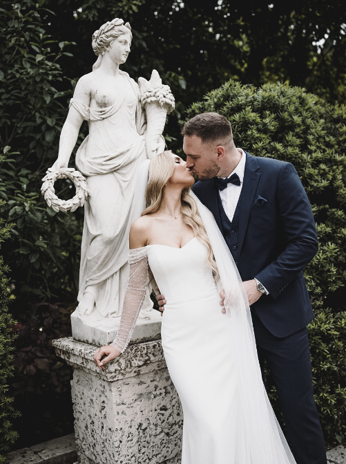 A bride in a white wedding gown and a groom in a navy suit kissing in front of a stone statue in a garden.