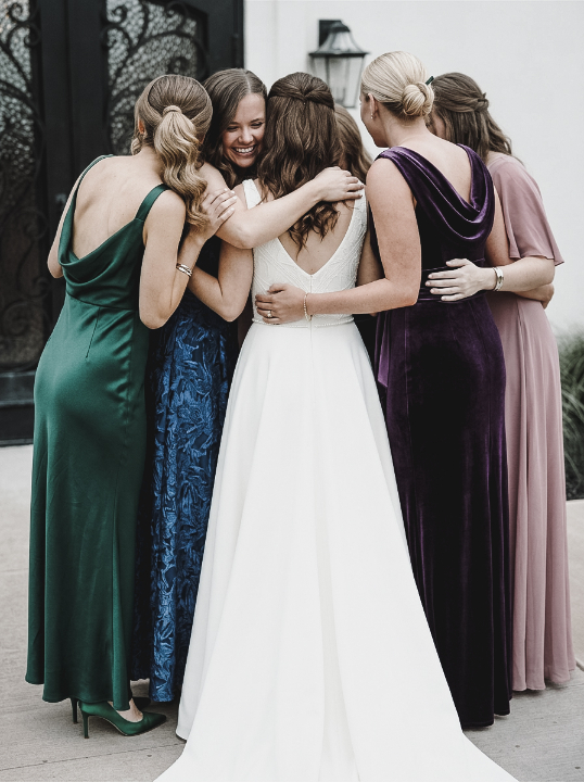 A bride in a white gown is hugged by five bridesmaids wearing long dresses in various colors.