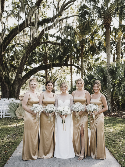 A bride in a white wedding dress standing with four bridesmaids in matching gold gowns outdoors under oak trees.