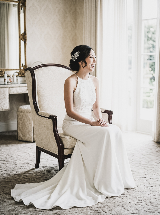 A bride in a white halter wedding dress with a beaded bodice laughs while seated in a chair.