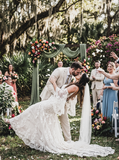 A groom dips his bride for a kiss at their outdoor wedding, surrounded by guests and flower petals in the air.