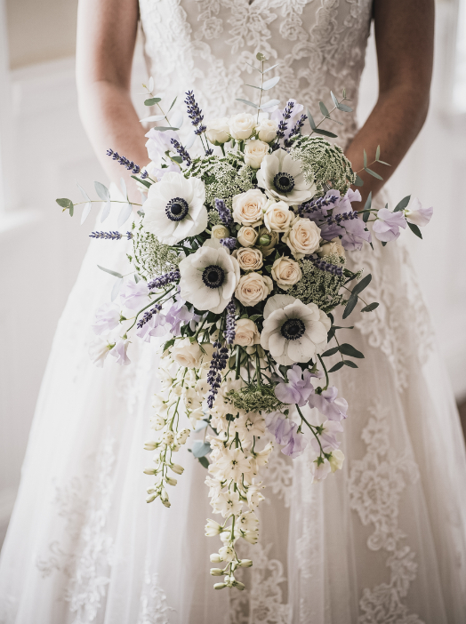 A bride in a lace wedding dress holds a cascading bouquet of white anemones, pale roses, lavender, and greenery.