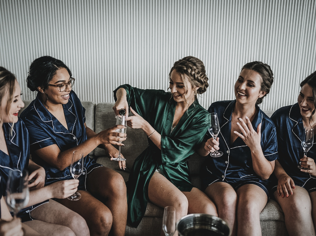 Five women in pajamas and robes sitting on a couch, with one woman pouring champagne into a glass.