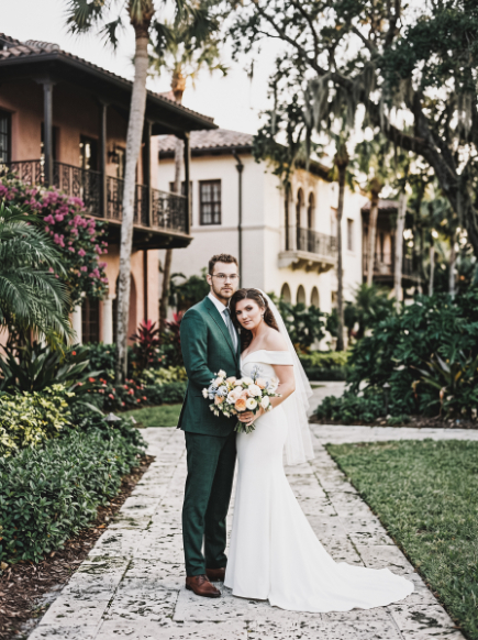 A bride in a white wedding gown and a groom in a dark green suit stand together holding a bouquet on a stone path in front of a mediterranean-style building.