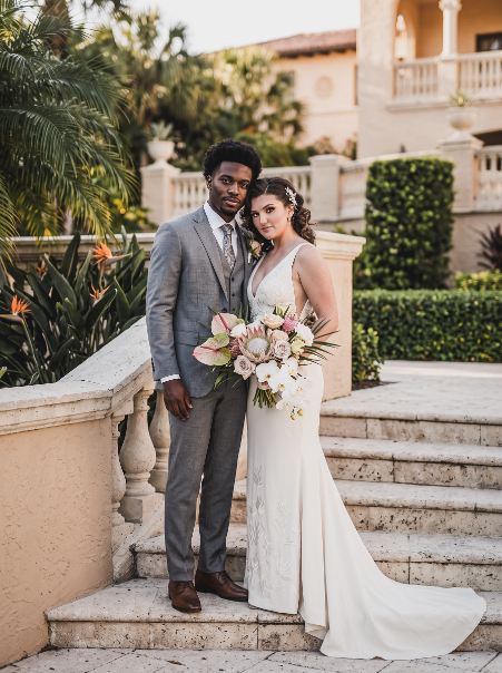 A bride in a white wedding gown and a groom in a grey suit stand together on stone stairs, holding a large floral bouquet.