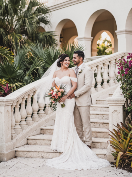 A bride in a white wedding gown and a groom in a beige suit stand together on a stone staircase.