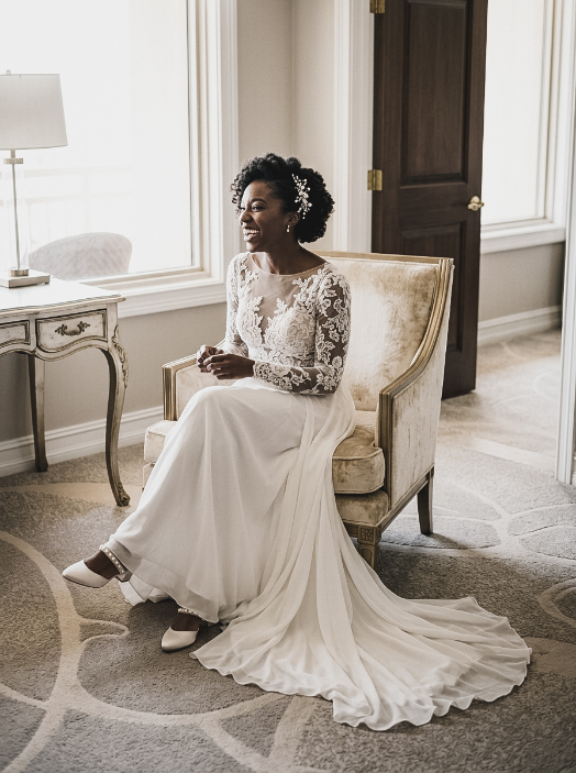 A smiling bride in a long-sleeved lace wedding dress sitting in an upholstered armchair.