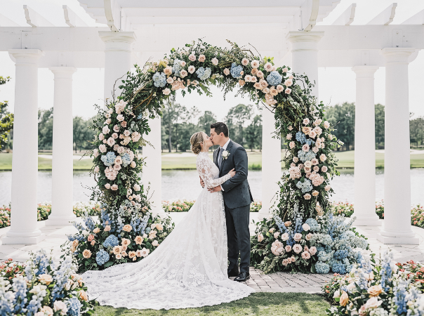 A bride and groom kissing under a large, circular floral archway between white pillars at an outdoor wedding.
