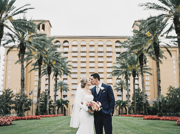 A bride and groom in wedding attire kiss on a lawn in front of a large hotel flanked by palm trees.