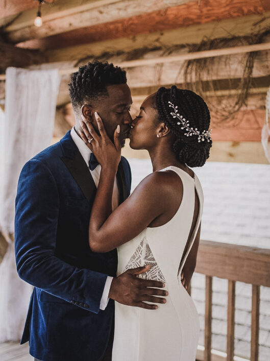 A bride in a white gown and a groom in a blue velvet tuxedo kiss on a wooden balcony.