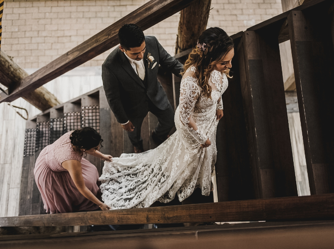 A bridesmaid in a mauve dress holds the long lace train of a bride’s gown as she and a groom in a dark suit walk up wooden stairs.