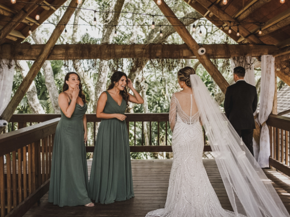 A bride in a beaded gown with a long veil stands before her groom while two bridesmaids look on with surprised expressions.