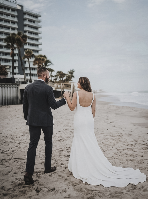 A bride in a white wedding gown and a groom in a dark suit walk hand-in-hand along a sandy beach near a large hotel.