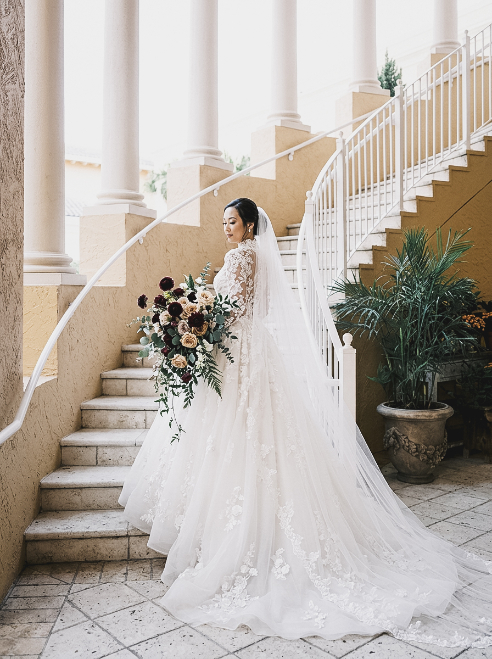 A bride in a long-sleeved lace wedding gown and veil stands on stone stairs holding a large bouquet of dark red and white flowers.